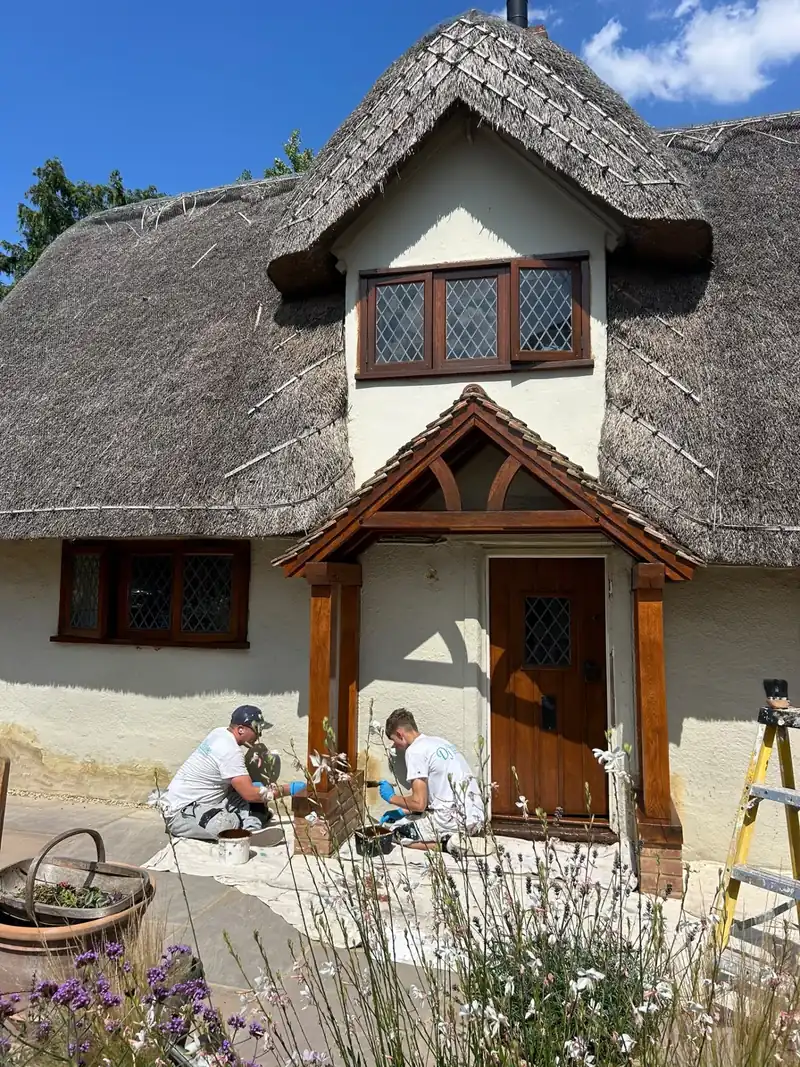 Two painters at work on the fresh render and oak porch of a thatched cottage